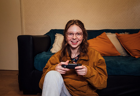 A gamer sitting on the floor resting on the soft. She's staring directly to the camera smiling.)