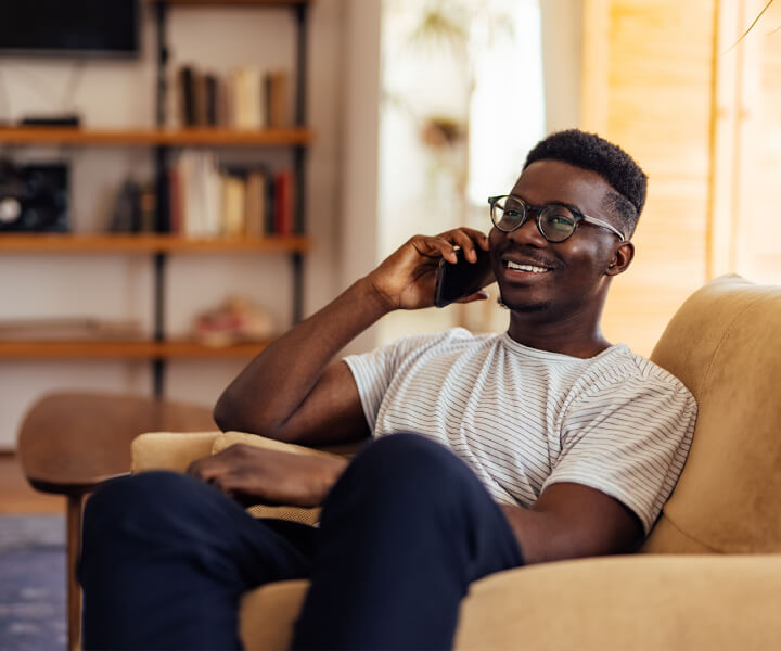 A man sitting on a chair, on his phone smiling. He appears to be in a living room, with bookshelves out of focus behind him.