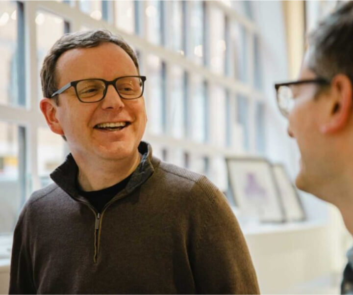 A man in glasses looking away from the camera smiling at another man. The second man is out of focus.)
