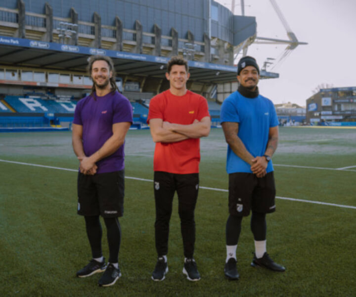 Three rugby players standing with their arms crossed. The Cardiff Arms Park stadium can be seen in the background.