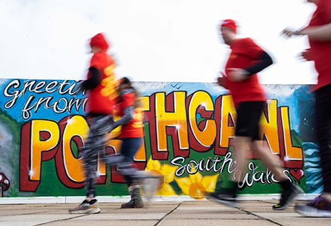 Four runners out of focus running passed a sign that reads 'Greetings from Porthcawl, south Wales'.)