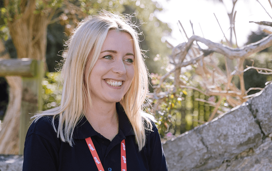 A woman looking away from the camera smiling. A wall and greenery can be seen behind her suggesting she's in a community garden setting.