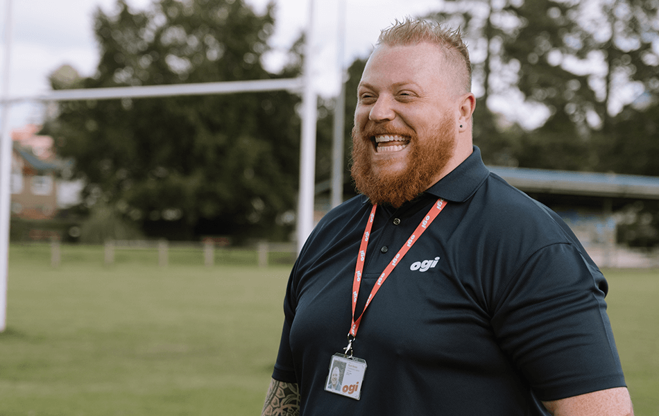 A man in an Ogi uniform looking away from the camera smiling. A rugby pitch with posts and a spectators stand can be seen out of focus behind him.