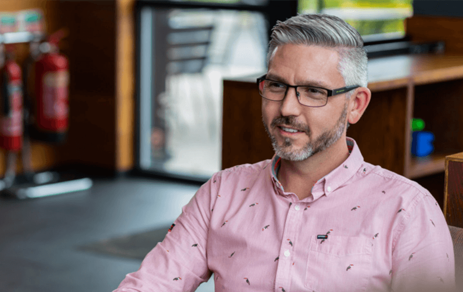 A man in glasses a pink shirt looking away from the camera smiling.