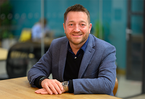 A man sitting at a table hands crossed looking to the camera smiling.)