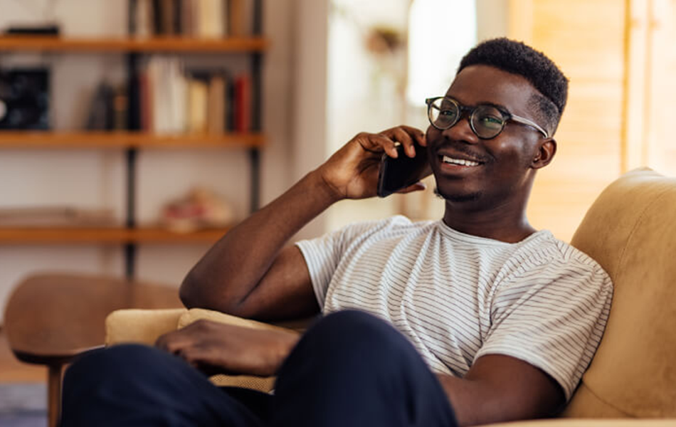 A man sitting on a chair, on his phone smiling. He appears to be in a living room, with bookshelves out of focus behind him.