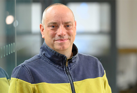 A man in a sweatshirt looking to the camera. A modern office can be seen behind him slightly out of focus.)