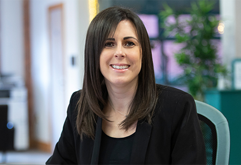 A woman in a suit looking to the camera smiling. Out of focus you can see a modern office with plants and a printer.)