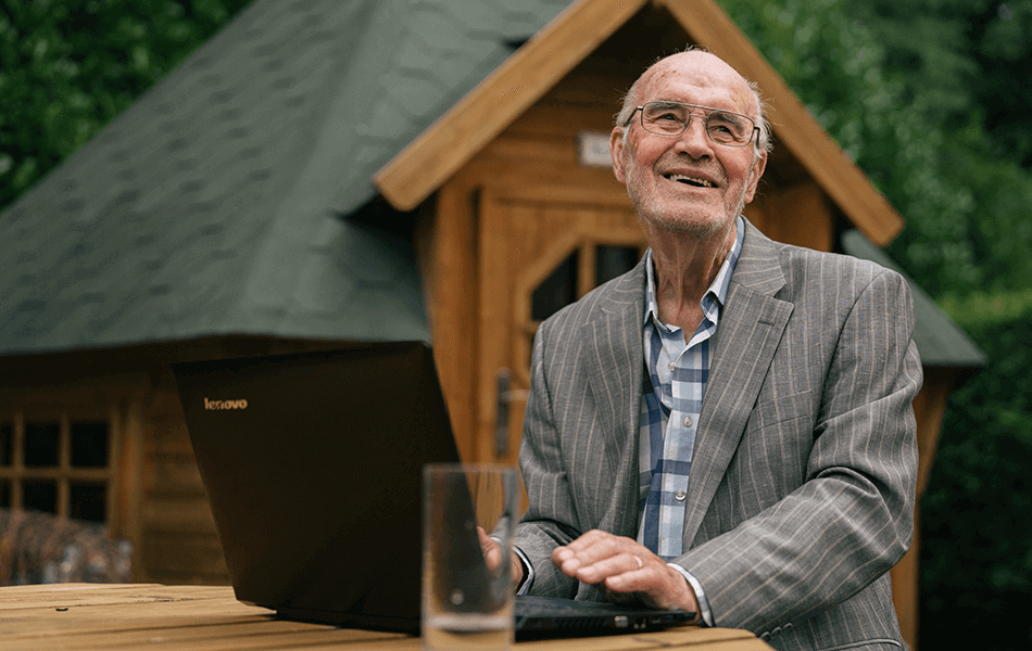 An older man looking away from the camera smiling. He's on a laptop in the garden with a large summer house slightly out of focus behind him.