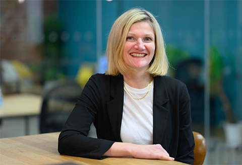 A woman sitting at a table leaning in smiling.)