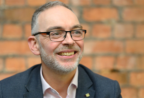 A man in glasses and a suit looking away from the camera smiling.)