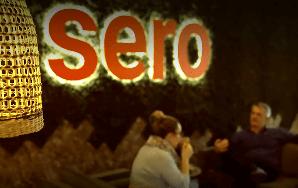 Two people sitting on a sofa chatting. A large illuminated sign spelling 'Sero' can be seen in focus.