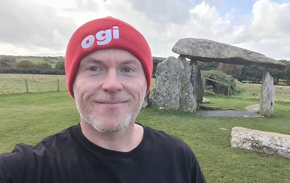 A man smiling at the camera in an Ogi beanie. An ancient stone monument and rolling countryside hills can be seen in the background.