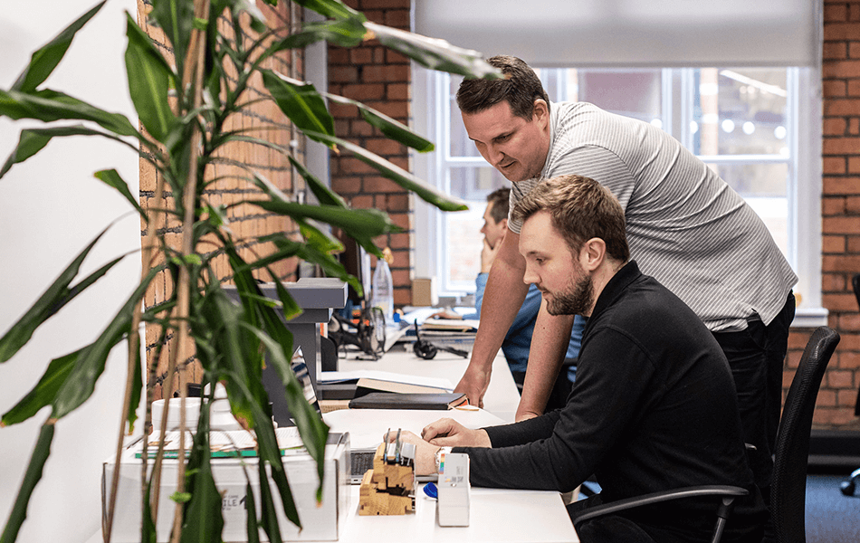 Two people in an office. One is leaning over the offer discussing something. They both appear engrossed in the work.