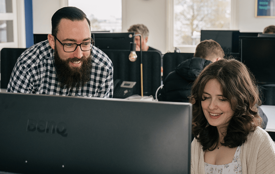 Two people looking at a computer screen smiling. A busy office can be seen behind them.