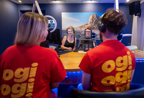 Four in a radio studio. Two have their backs to the camera with bright t-shirts on with the words 