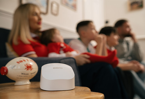 A close up of an eero router next to a ceramic Welsh rugby ball. A family can be seen blurred out in the background.)