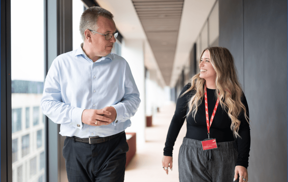 Two people walking along a corridor chatting. They are smiling and appear engaged in conversation. The corridor is in a modern office with floor to ceiling glass to one side. You can just about see the city scape through the windows.