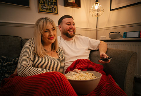 Couple sitting on the sofa in a dimply lit living room. With a blanket over them and a big bowl of popcorn perched, they are ready to watch a film.)
