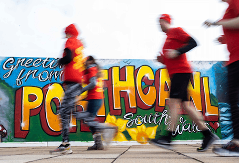 Four runners out of focus running passed a sign that reads 'Greetings from Porthcawl, south Wales'.)