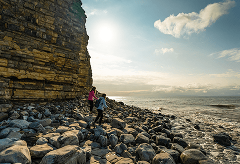 Two people walking along the pebbles of the Vale of Glamorgan coastline. A choppy sea and moody sky can be seen in the distance.